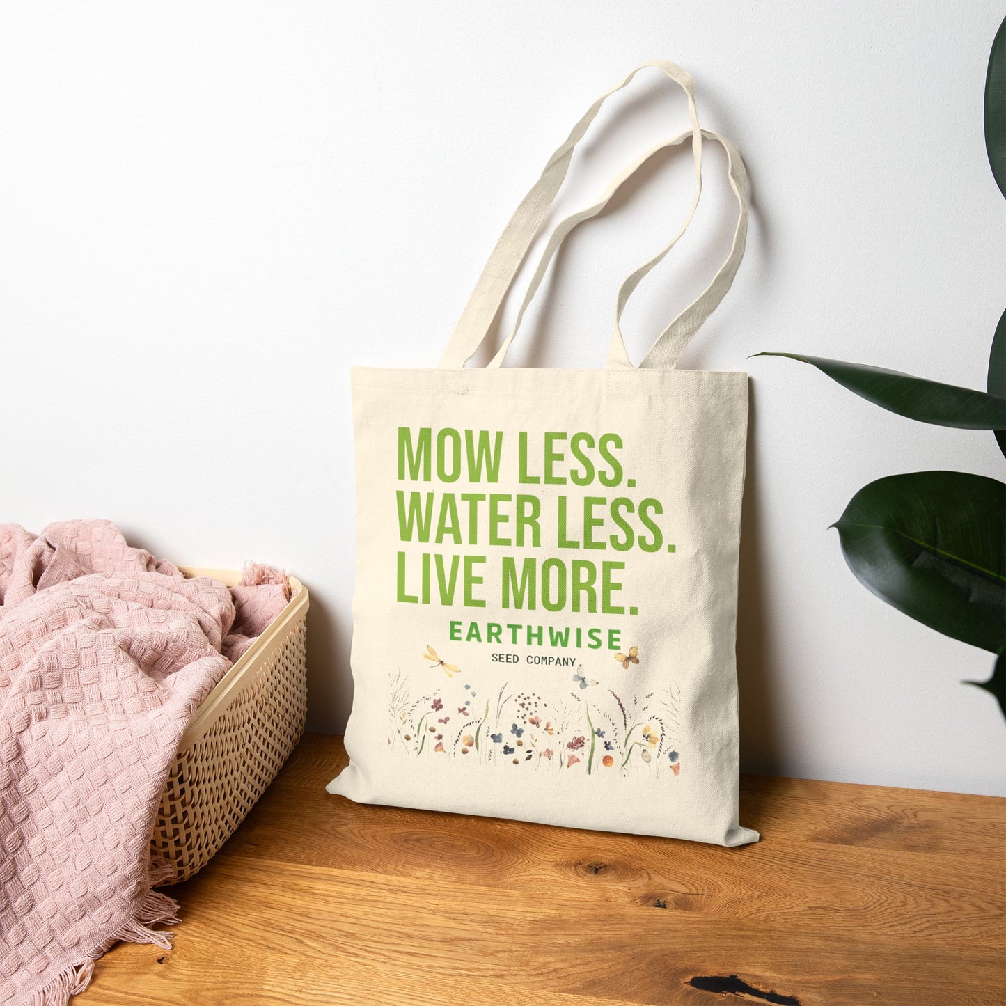 Tote bag with 'Mow Less, Water Less, Live More' text on a wooden surface with a plant in the background