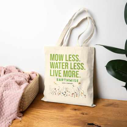Tote bag with 'Mow Less, Water Less, Live More' text on a wooden surface with a plant in the background
